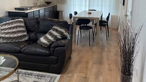 Moderni olohuone, jossa on musta nahkasohva, ruokapöytä tuoleineen, kattotuuletin sekä keittiöalue, jossa on pesuallas ja kaappeja.