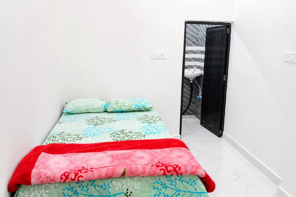 Quarto pequeno e bem arrumado, com cama, travesseiros e um banheiro visível por uma porta aberta.