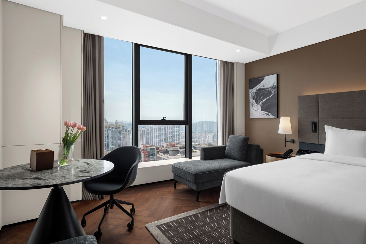 A hotel room with a bed, a desk, a chair, a view of the city, and a painting on the wall.