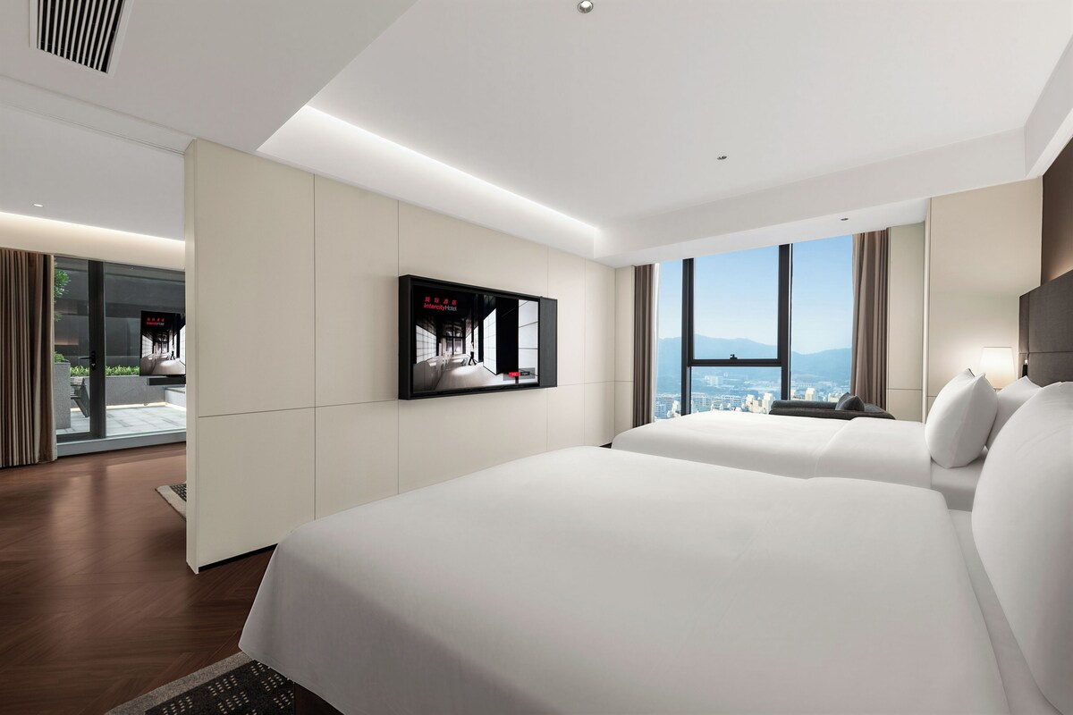 A modern hotel room with two beds, a TV mounted on the wall, and a large window offering a city view.