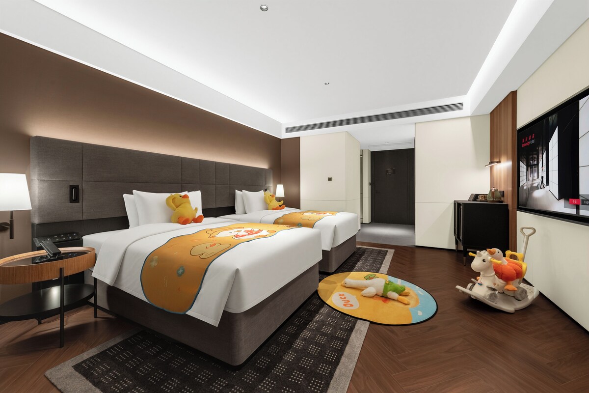 A hotel room with two beds, a desk, a lamp, a TV, and a small table with toys.