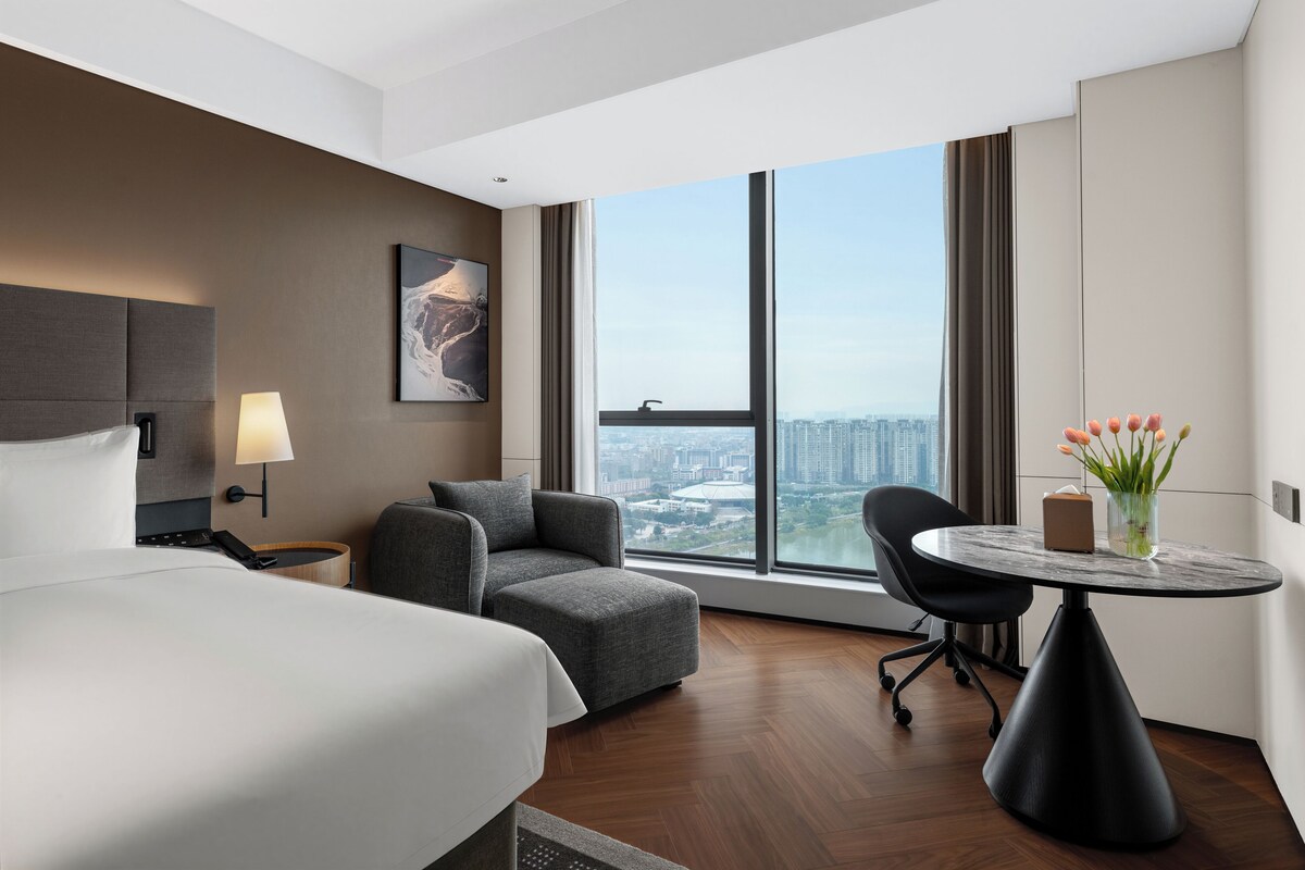 A hotel room with a bed, a sofa, a desk, and a large window offering a city view.