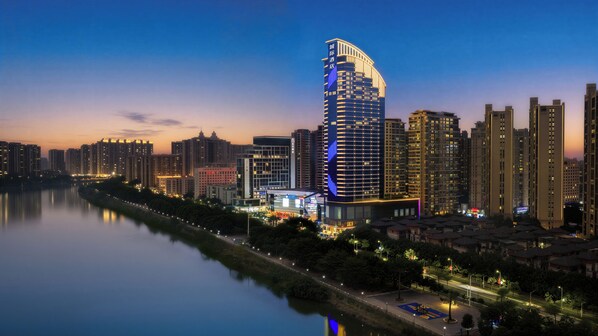 A modern high-rise hotel with a curved facade, located near a river with a well-lit promenade.