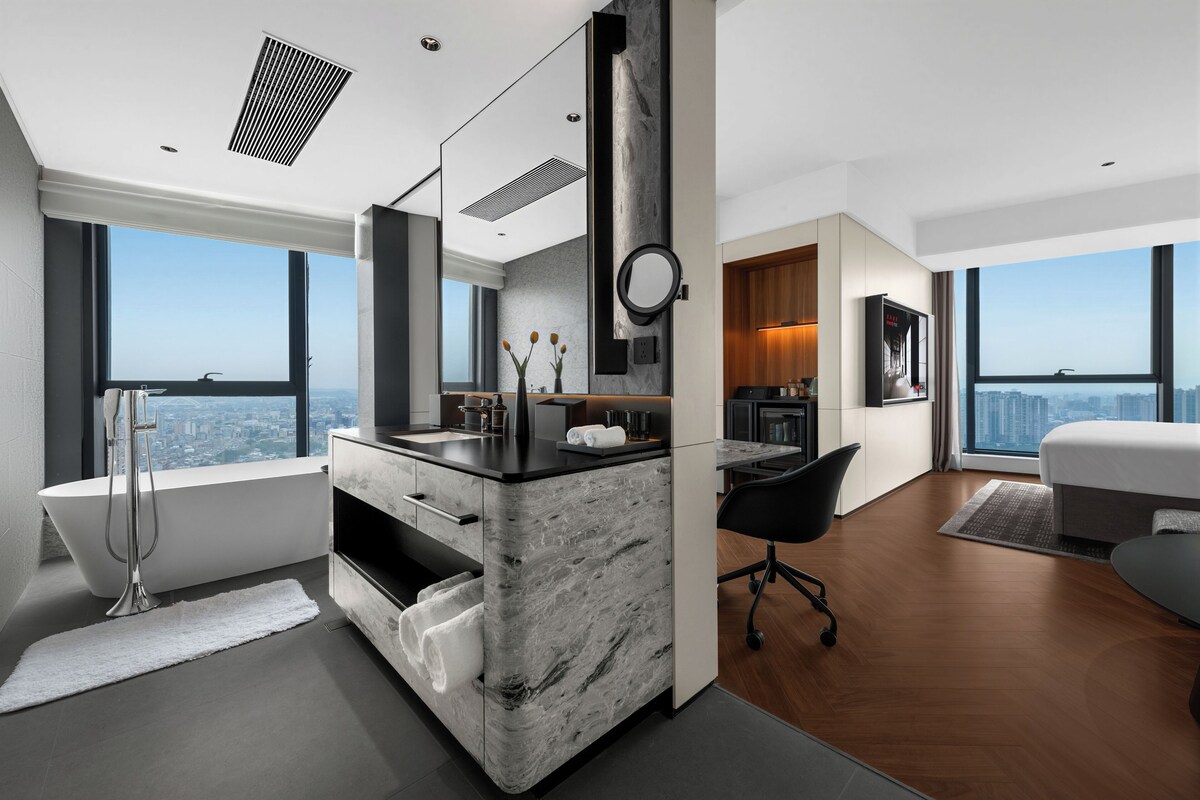 A modern hotel room with a large window, a marble bathroom vanity, a black office chair, and a bed.