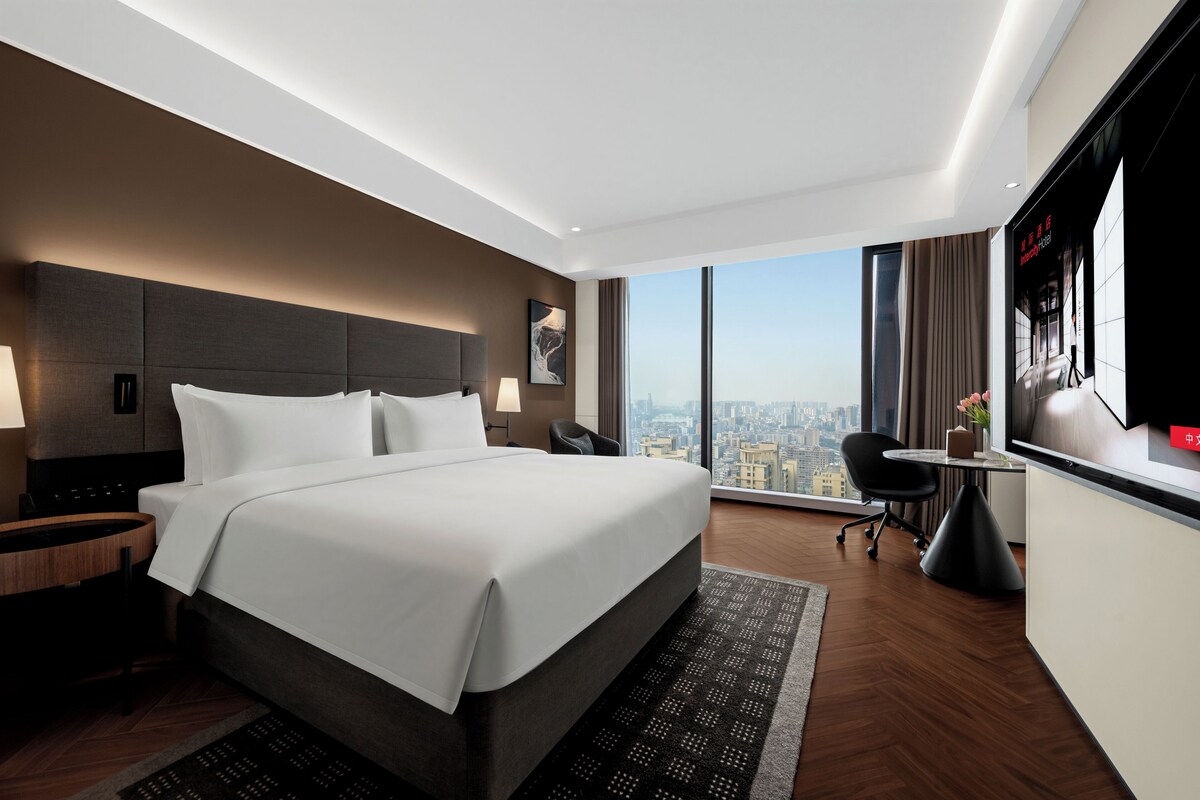 A modern hotel room with a large bed, a desk, a chair, and a city view.