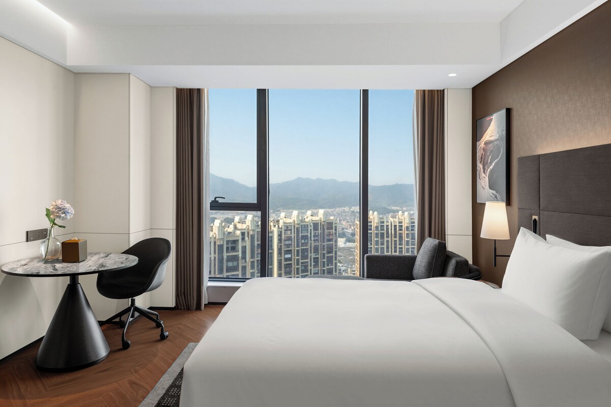 A hotel room with a large bed, a desk with a chair, a view of the city, and a window with curtains.