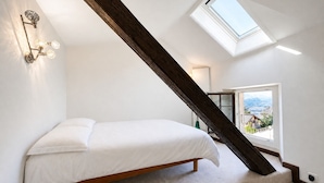 A bedroom with a sloped ceiling, a skylight, a bed, and a view of a landscape through a window. - Open Sky Retreat Panoramic Alpine Views 6 Sleeps (Chelin)