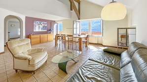 A spacious living area with a high ceiling, a dining table, a sofa, and a large window offering a view. - Open Sky Retreat Panoramic Alpine Views 6 Sleeps (Chelin)