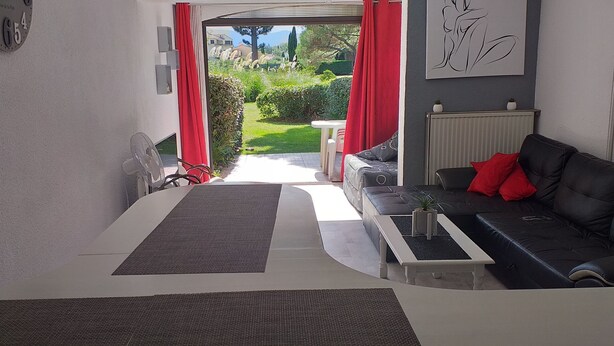 A modern living room with a sofa, a coffee table, a fan, and a view of a garden through a glass door. - Beautiful apartment in a large wooded park (Saint-Cyprien)