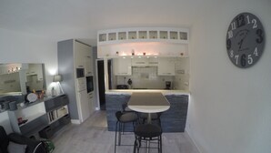 A modern kitchen with a central island, a dining area with two chairs, and a large wall clock. - Beautiful apartment in a large wooded park (Saint-Cyprien)