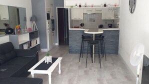 A modern living room with a black sofa, a white coffee table, and a dining area with a small island and black chairs. - Beautiful apartment in a large wooded park (Saint-Cyprien)