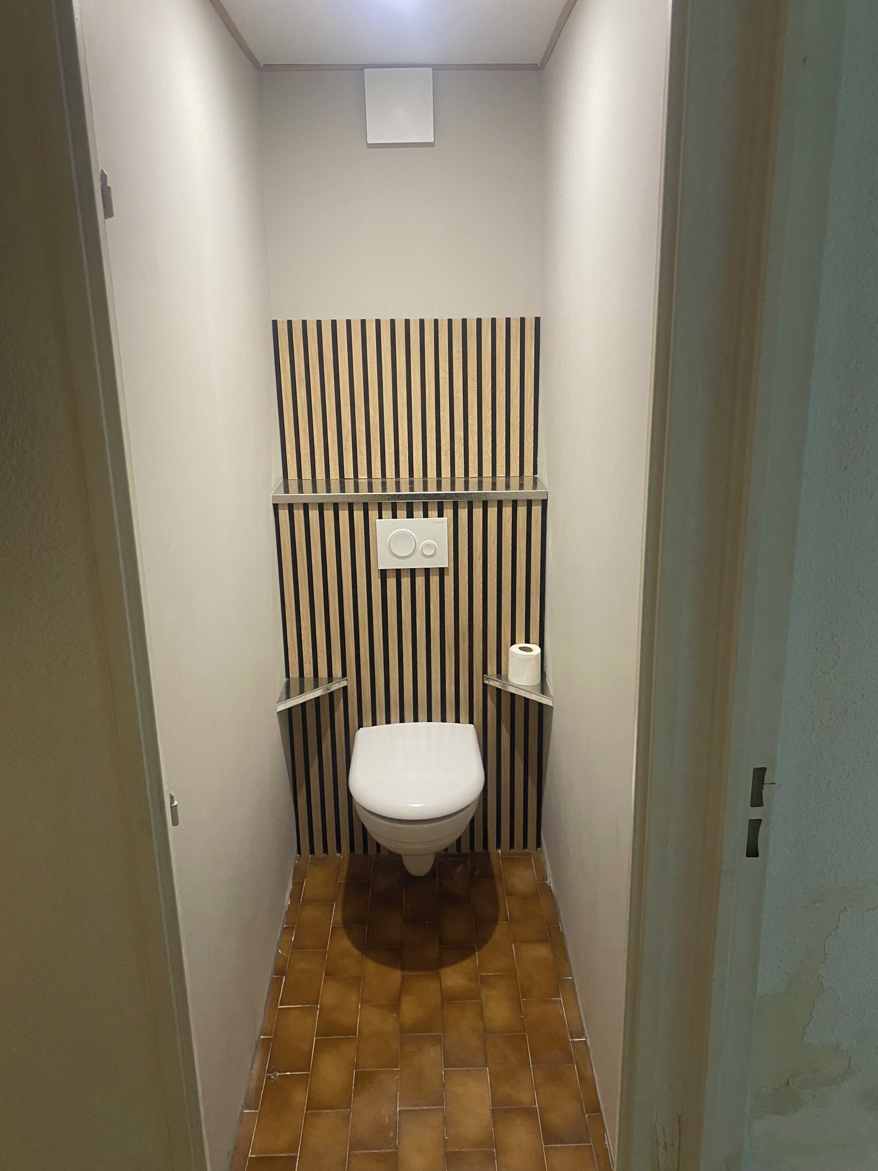 Une petite salle de bain comprenant des toilettes, une étagère avec un rouleau de papier toilette et un luminaire fixé au mur.