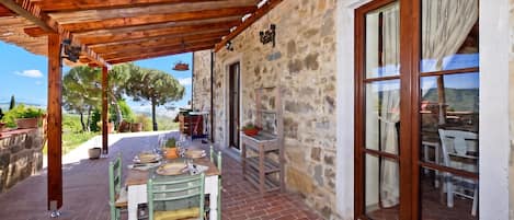 A covered outdoor dining area with a table set for two, stone walls, and a view of the countryside.