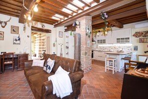A cozy living room with a brown leather sofa, a kitchen area with a brick wall, and a dining space with wooden furniture. - Dimora Belfiore (Capolona)