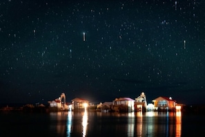 En ejendom ved vandet med oplyste bygninger, der spejles i vandet, under en stjernetyk himmel.