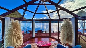 Een kamer met uitzicht op een meer, een rieten dak en een houten terras.