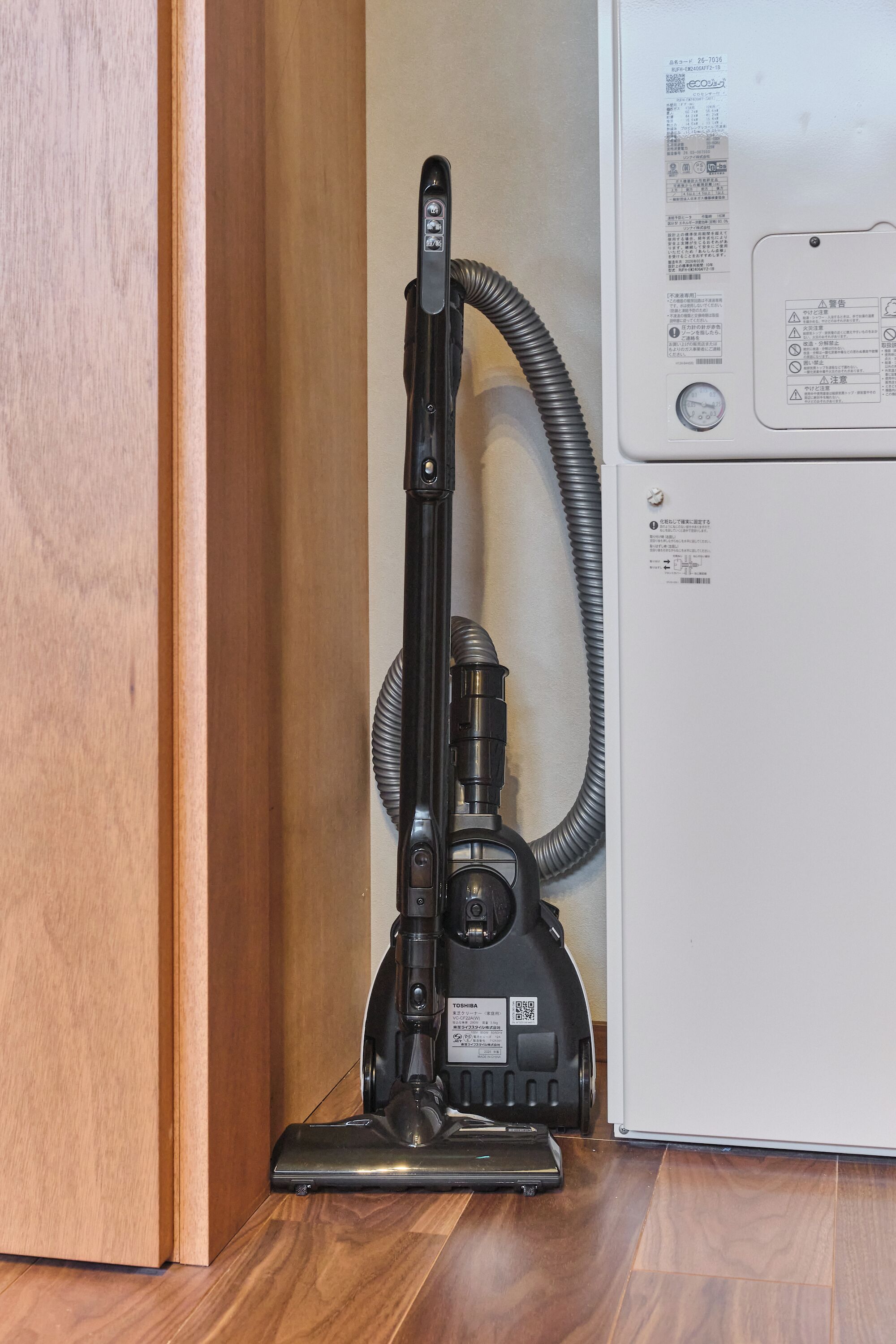 Een stofzuiger naast een wit apparaat, mogelijk een koelkast, in een kamer met houten vloeren.