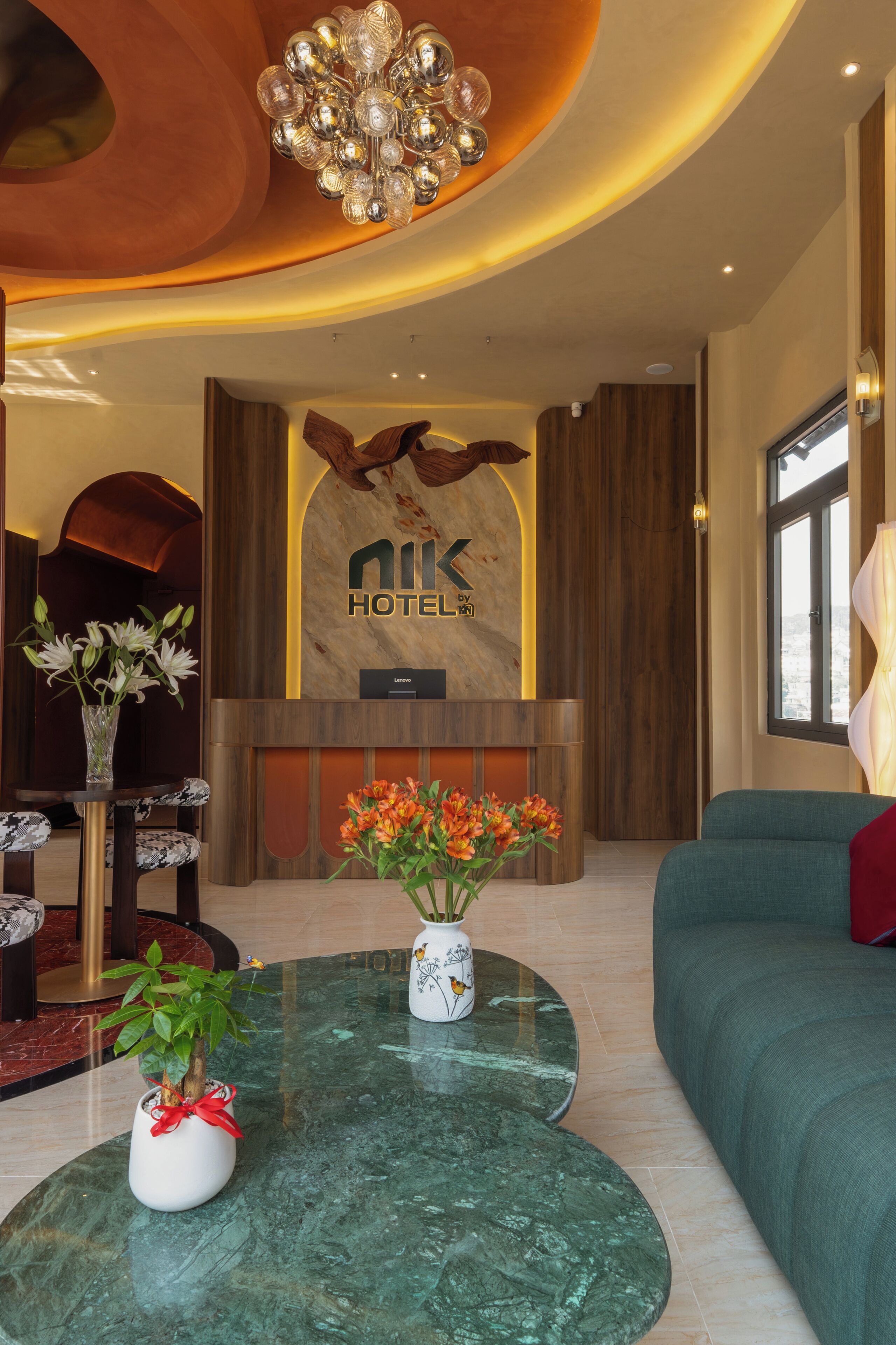 A hotel reception area with a reception desk, a large chandelier, a sofa, and two vases with flowers.