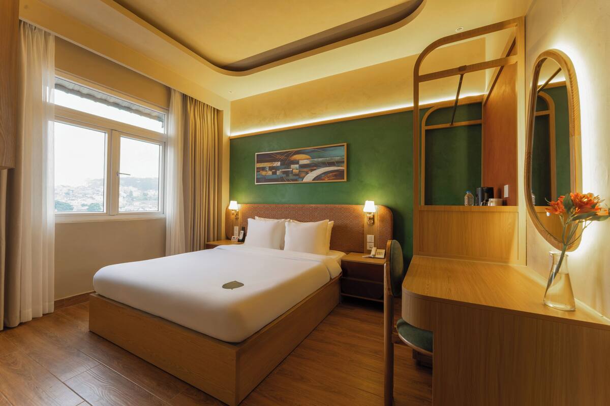 A hotel room with a bed, a desk, a chair, a mirror, a nightstand, a painting on the wall, and a window with curtains.