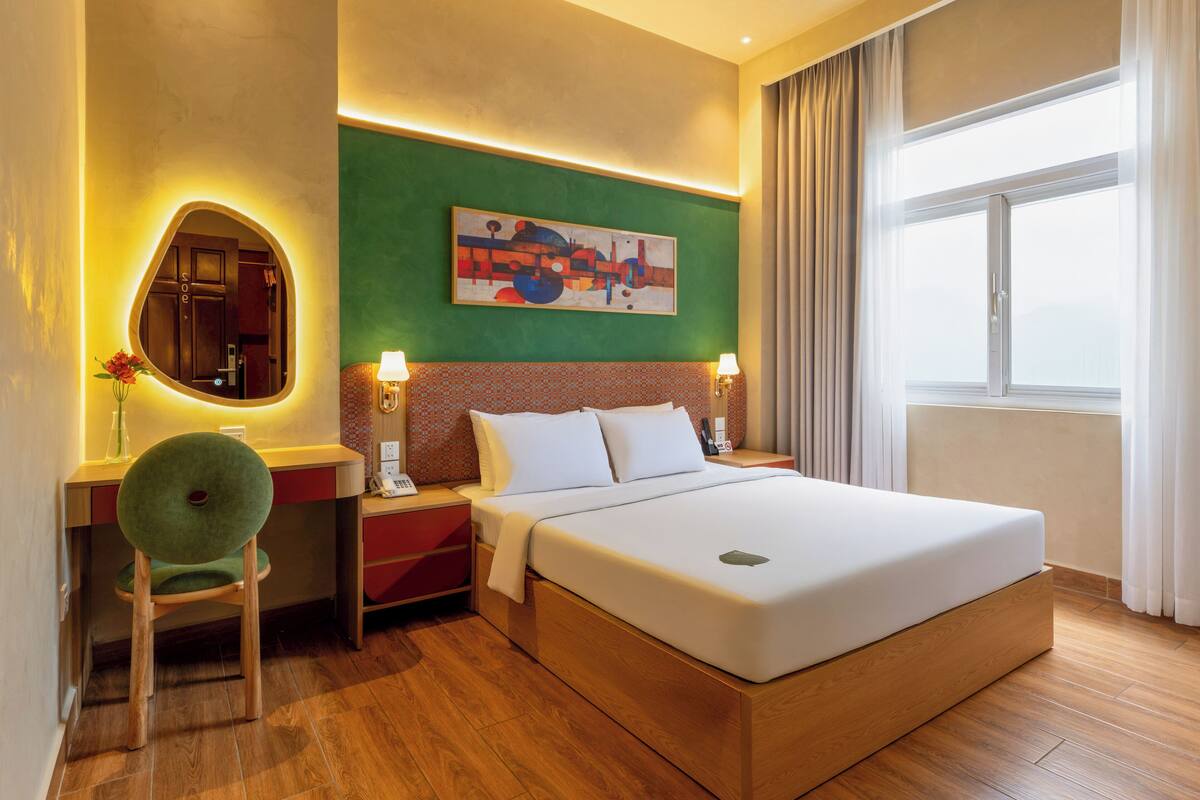 A hotel room with a bed, a desk, a chair, a painting on the wall, and a window with curtains.