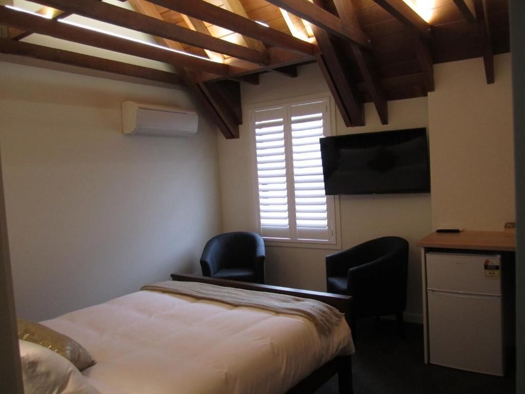 A bedroom with a bed, two chairs, a desk, a television, and a window with blinds.