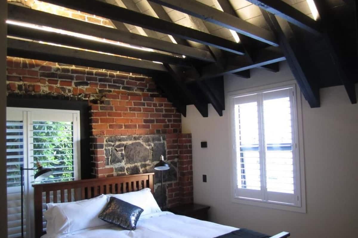 Un dormitorio con pared de ladrillo, cama de madera, ventana con persianas y techo con vigas a la vista.