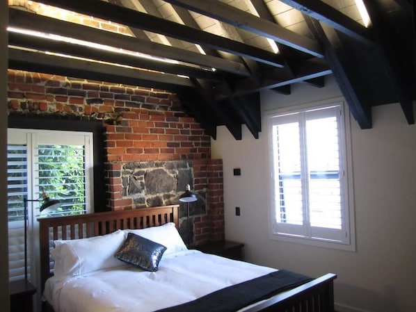 A bedroom with a brick wall, a wooden bed, a window with blinds, and a ceiling with exposed beams.