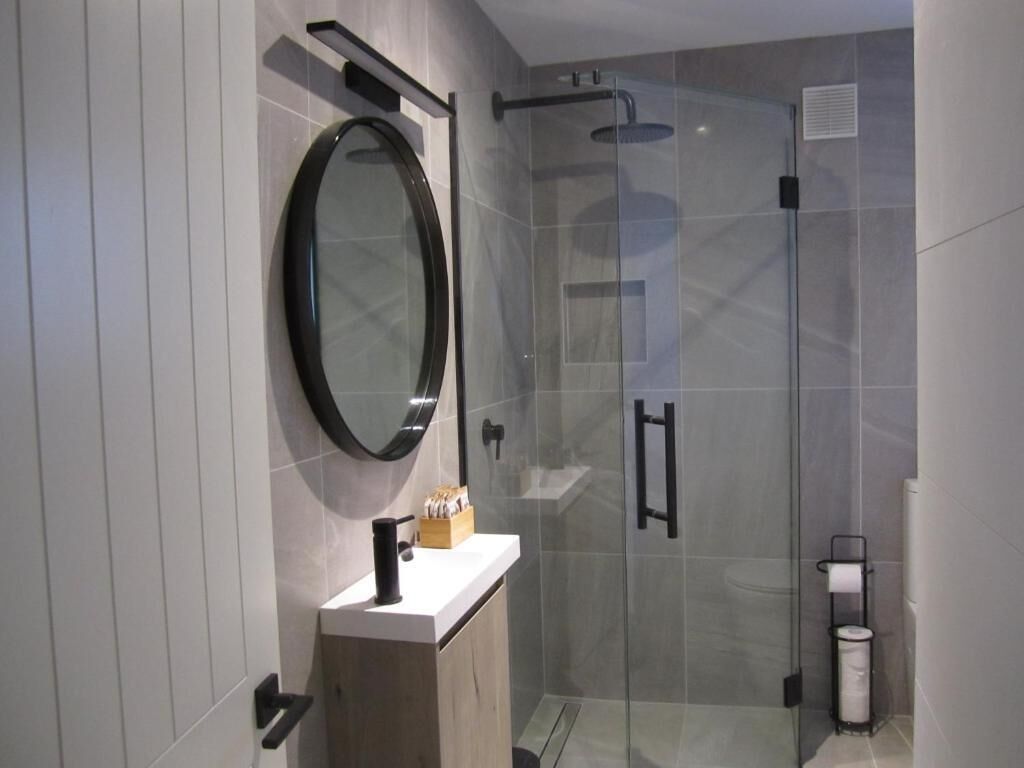 Un baño moderno con cabina de ducha de vidrio, un espejo redondo y un mueble de tocador de madera.