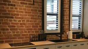Una cocina con pared de ladrillo, estufa negra, armarios blancos y una ventana con persianas.