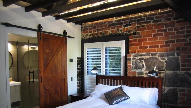 Un dormitorio con una cama, cabecera de madera, pared de piedra, ventana con persianas y una puerta corrediza de estilo rústico.