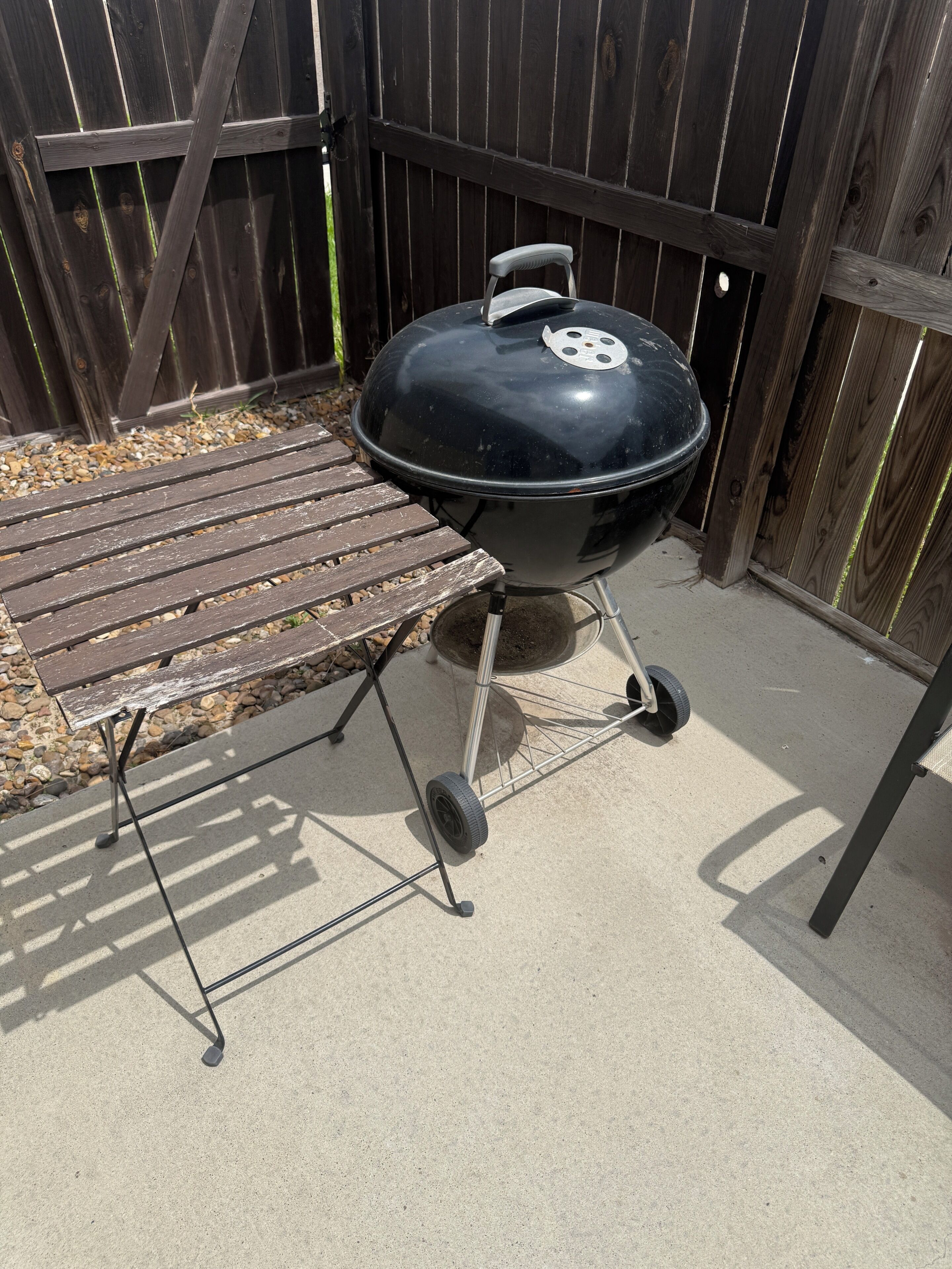 A black barbecue grill with wheels, a wooden bench, and a wooden fence in the background.