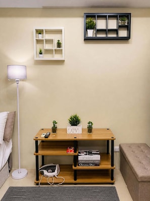 A room with a bed, a bedside lamp, a wooden console table with books and a remote control, a small shelf with plants, and a wall-mounted shelf with more plants.