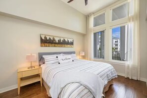 Ein Schlafzimmer mit einem Bett, einem Nachttisch, einer Lampe, einem Stadtbild als Gemälde und einem Fenster mit Vorhängen.