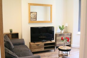 Ein Wohnzimmer mit Flachbildfernseher, einer Couch, einem Couchtisch, einem Beistelltisch mit Lampe und einer Vase mit Blumen.