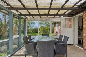 Une terrasse couverte avec une table en verre et des chaises en osier, un mur en briques et une porte donnant sur un jardin.