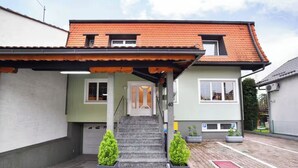 A two-story house with a red-tiled roof, a covered porch, and a staircase leading to the entrance. There are two potted plants on the ground floor.