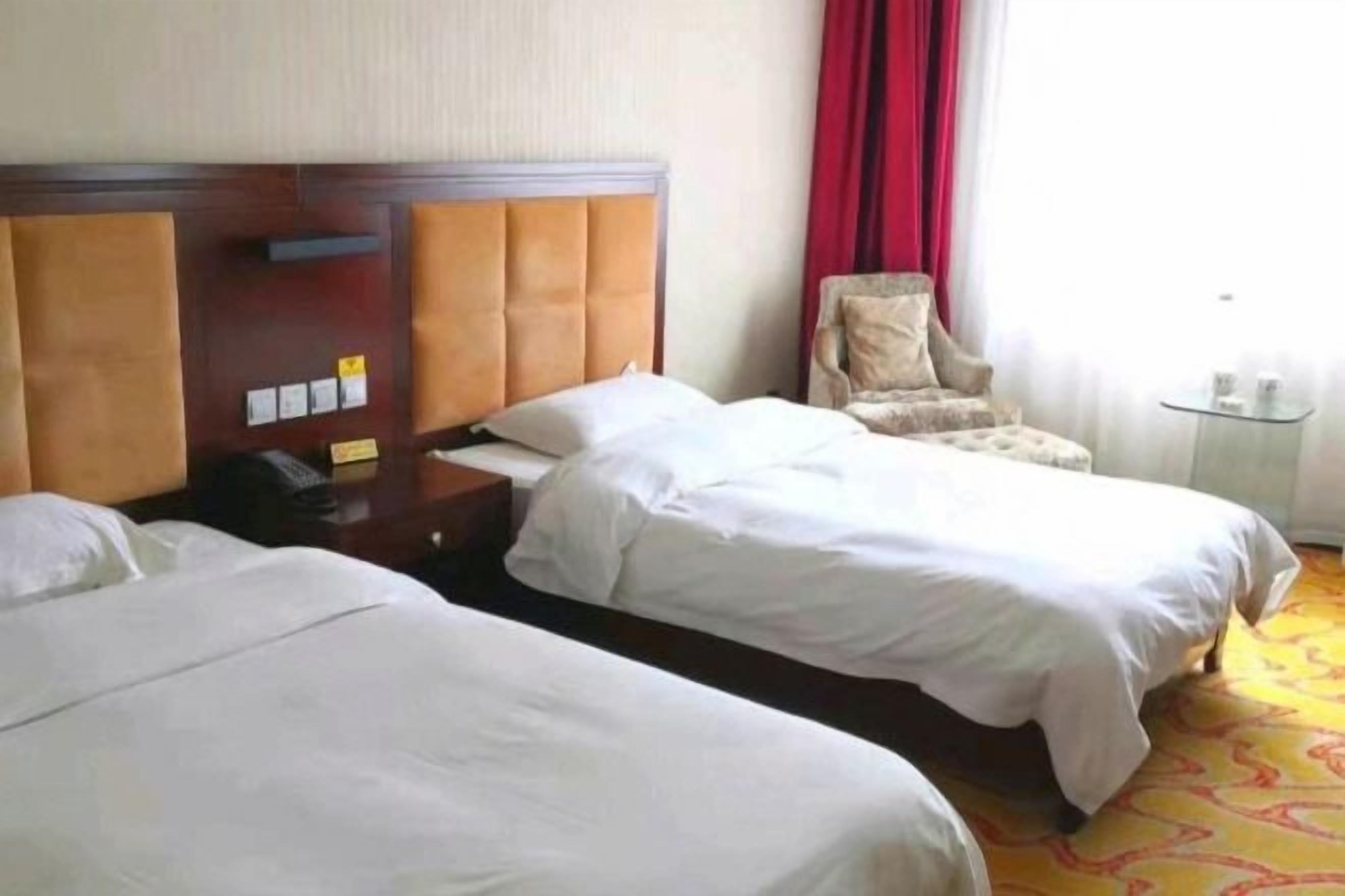 A hotel room with two beds, a wooden headboard, a chair, a bedside table, and a small table with a lamp.