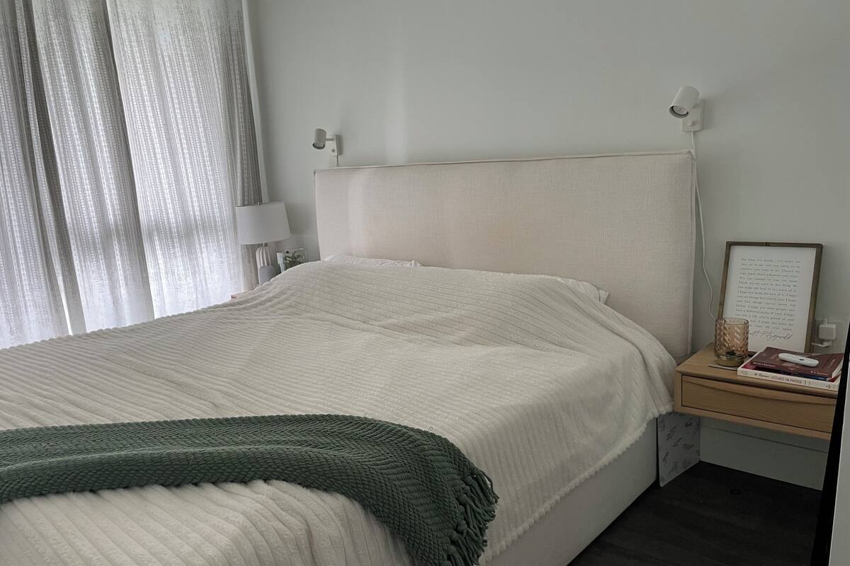 A neatly made bed with a green throw, a bedside table with a lamp, a framed picture, and a small plant.