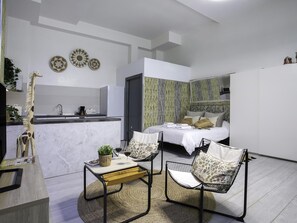 Quarto de hotel moderno com cozinha, cama com cabeceira, duas cadeiras, uma mesa pequena e arte decorativa na parede.