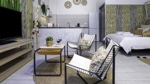 Una sala moderna con un televisor de pantalla plana, un comedor con una mesa pequeña y sillas, una cocina con refrigerador, y un dormitorio con cama y almohadas.