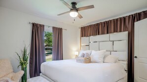 Ein Schlafzimmer mit einem großen Bett, einem Deckenventilator, einem Nachttisch, einem Sessel und einem Fenster mit Vorhängen.