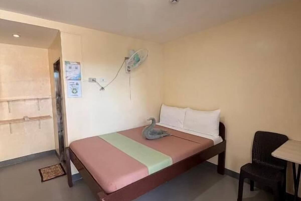 Una cama individual con almohada y manta, una mesita, una silla, un ventilador de pared y una puerta que da a otra habitación.