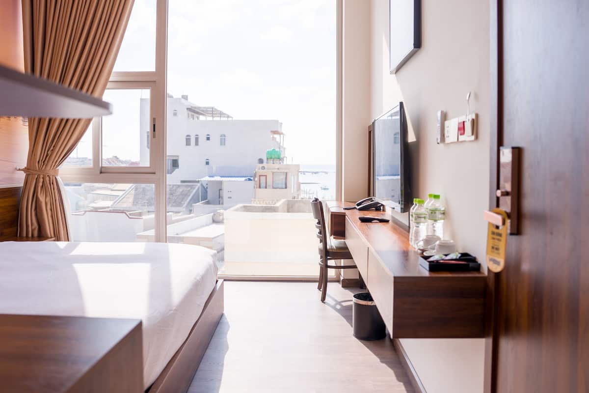 A hotel room with a bed, a desk, a television, and a view of buildings through a large window.