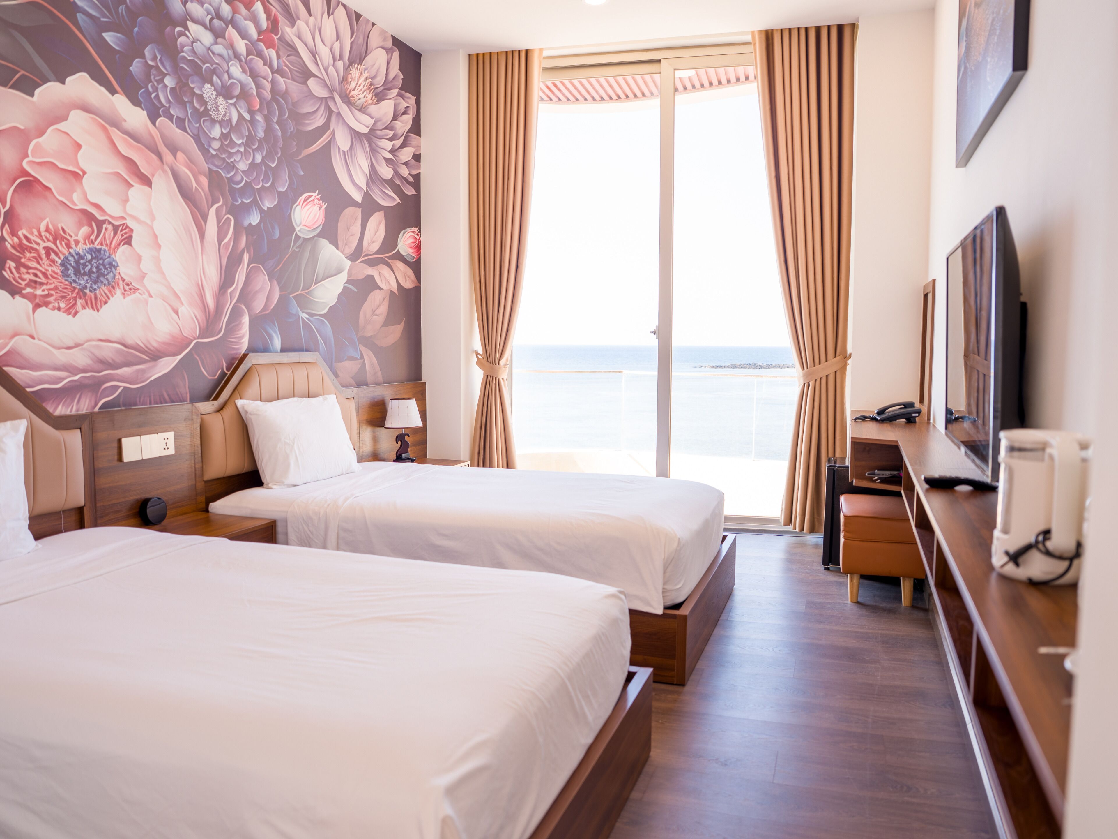 A hotel room with two beds, a desk, a TV, and a large floral mural.