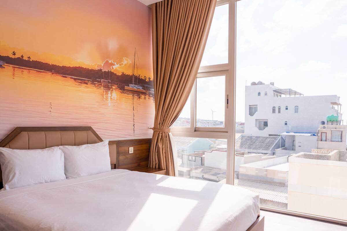 A hotel room with a bed, a large window, and a mural of a sunset over water.