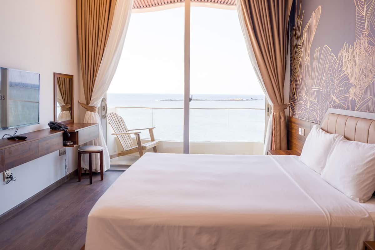 A hotel room with a bed, a desk with a telephone, a chair, and a view of the sea through a glass door.