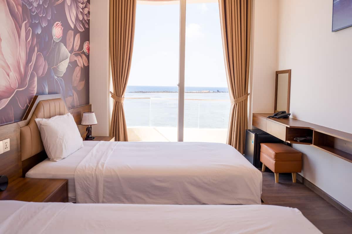 A hotel room with a bed, a desk, a chair, a lamp, a telephone, and a view of the sea.