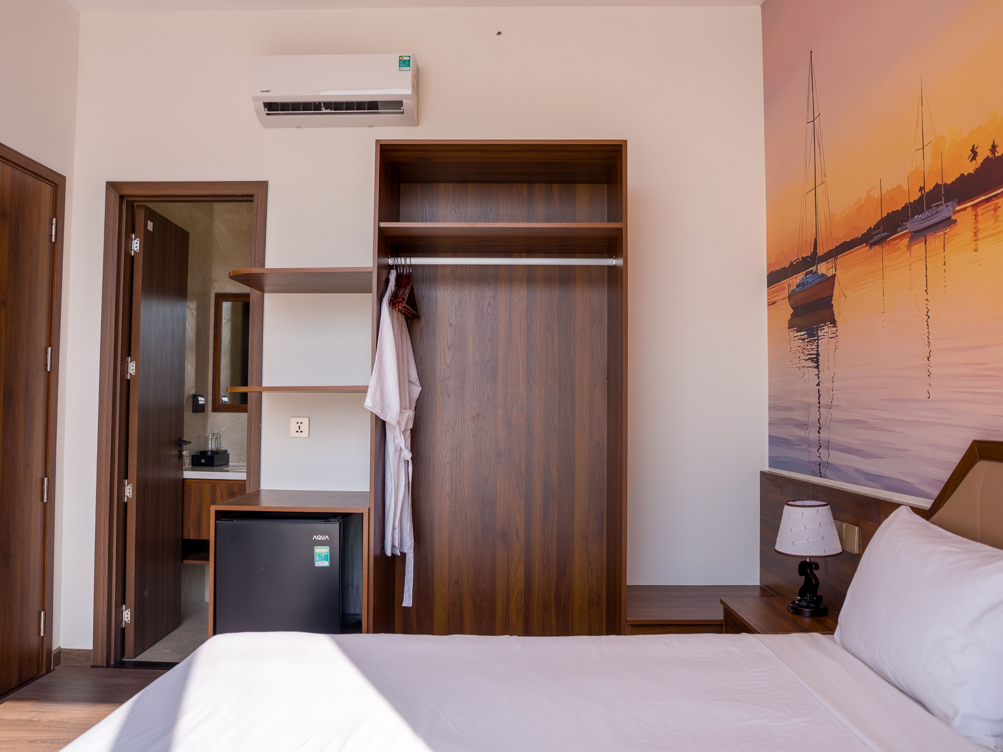 Ein Hotelzimmer mit Bett, Kleiderschrank, Schreibtisch, Kühlschrank, Wandaircondition und einem Gemälde von einem Sonnenuntergang über Wasser.