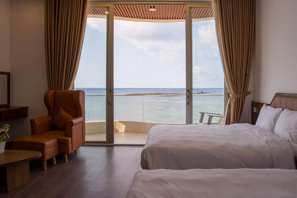 A hotel room with a bed, a chair, a small table, and a view of the sea through a glass door.