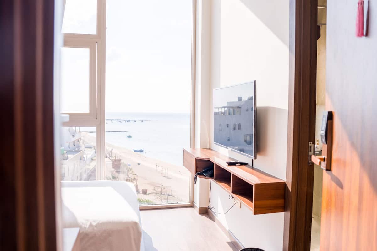 A room with a bed, a TV on a wooden shelf, a view of the sea, and a door.